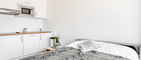 A modern bedroom with a bed, a bedside table with a plant, and a kitchenette with a microwave.