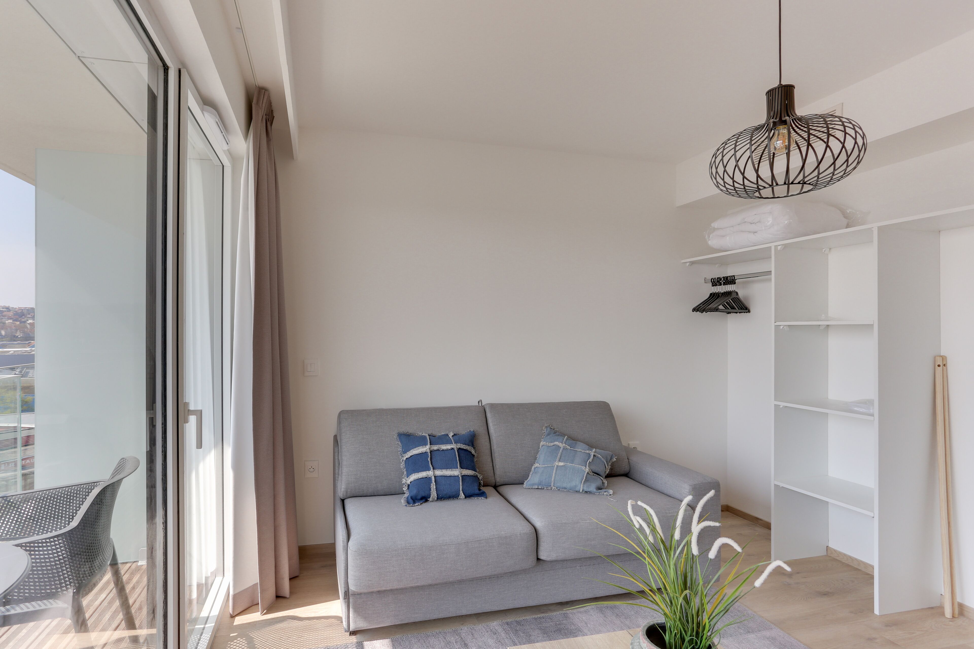 Una sala moderna con un sofá gris, una puerta de vidrio que da a un balcón y un mueble blanco con estantes.