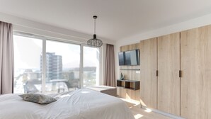 Een moderne slaapkamer met een groot bed, een aan een houten wand bevestigde televisie en een stadszicht door grote ramen.