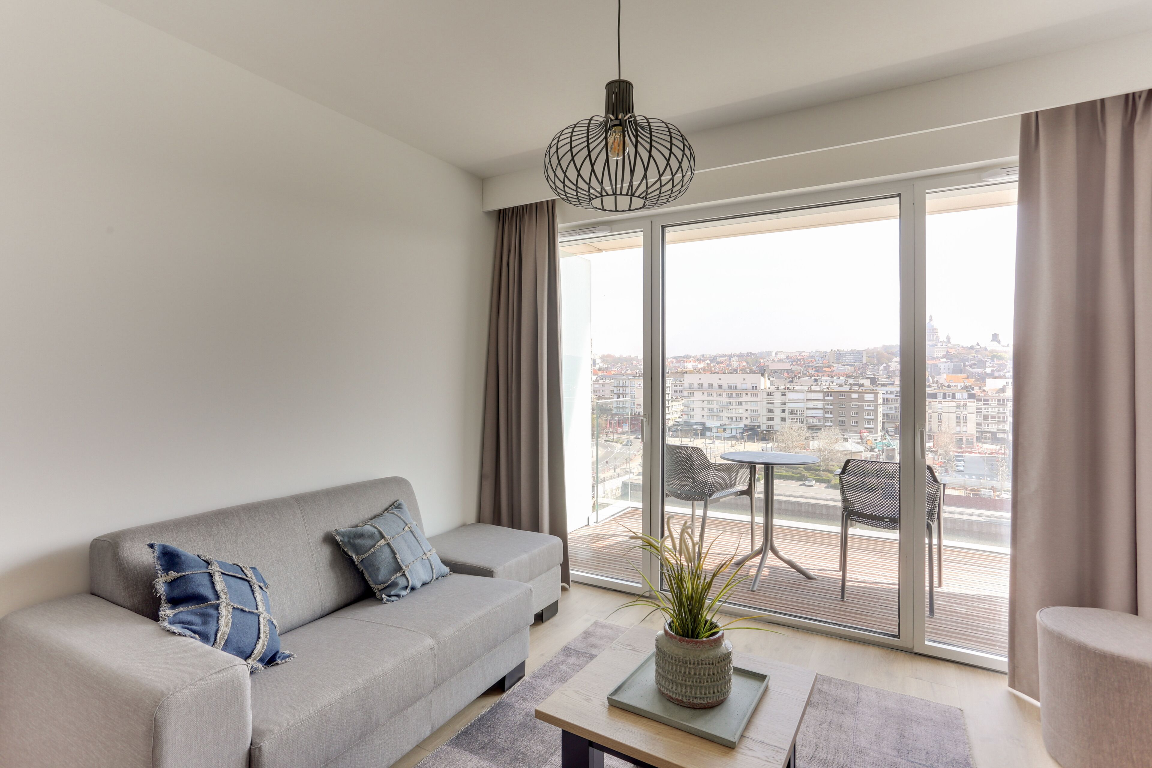 A modern living room with a grey sofa, a coffee table with a plant, and a city view through large windows.