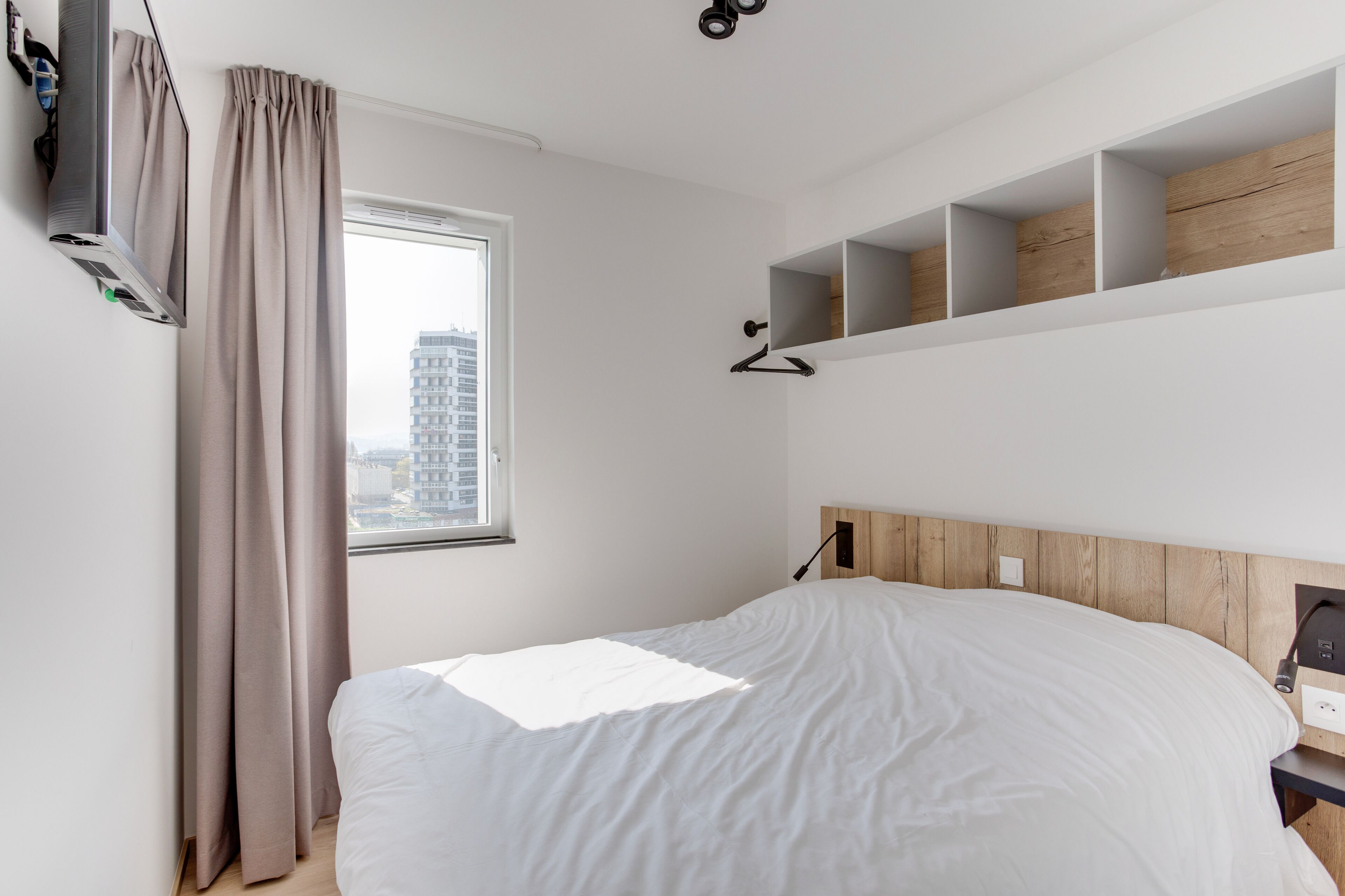 A modern hotel room with a bed, a window with curtains, a wall-mounted TV, and built-in shelves.