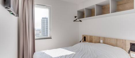 A modern hotel room with a bed, a window with curtains, a wall-mounted TV, and built-in shelves.