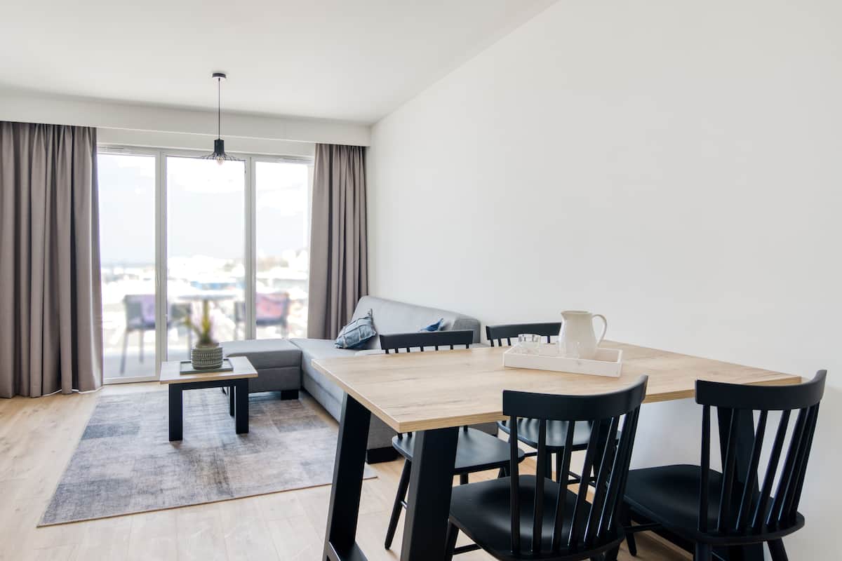 Ein modernes Wohnzimmer mit einem Holzesstisch, schwarzen Stühlen, einem grauen Sofa und einem großen Fenster mit Vorhängen.