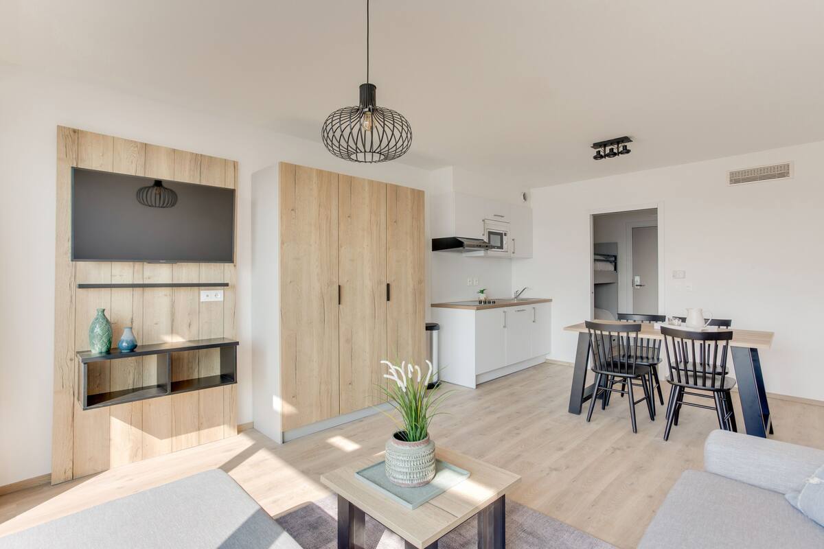 Ein modernes Wohnzimmer mit Holzboden, einer Essecke mit Tisch und Stühlen, einer Küche mit weißen Schränken und einem großen Kleiderschrank.
