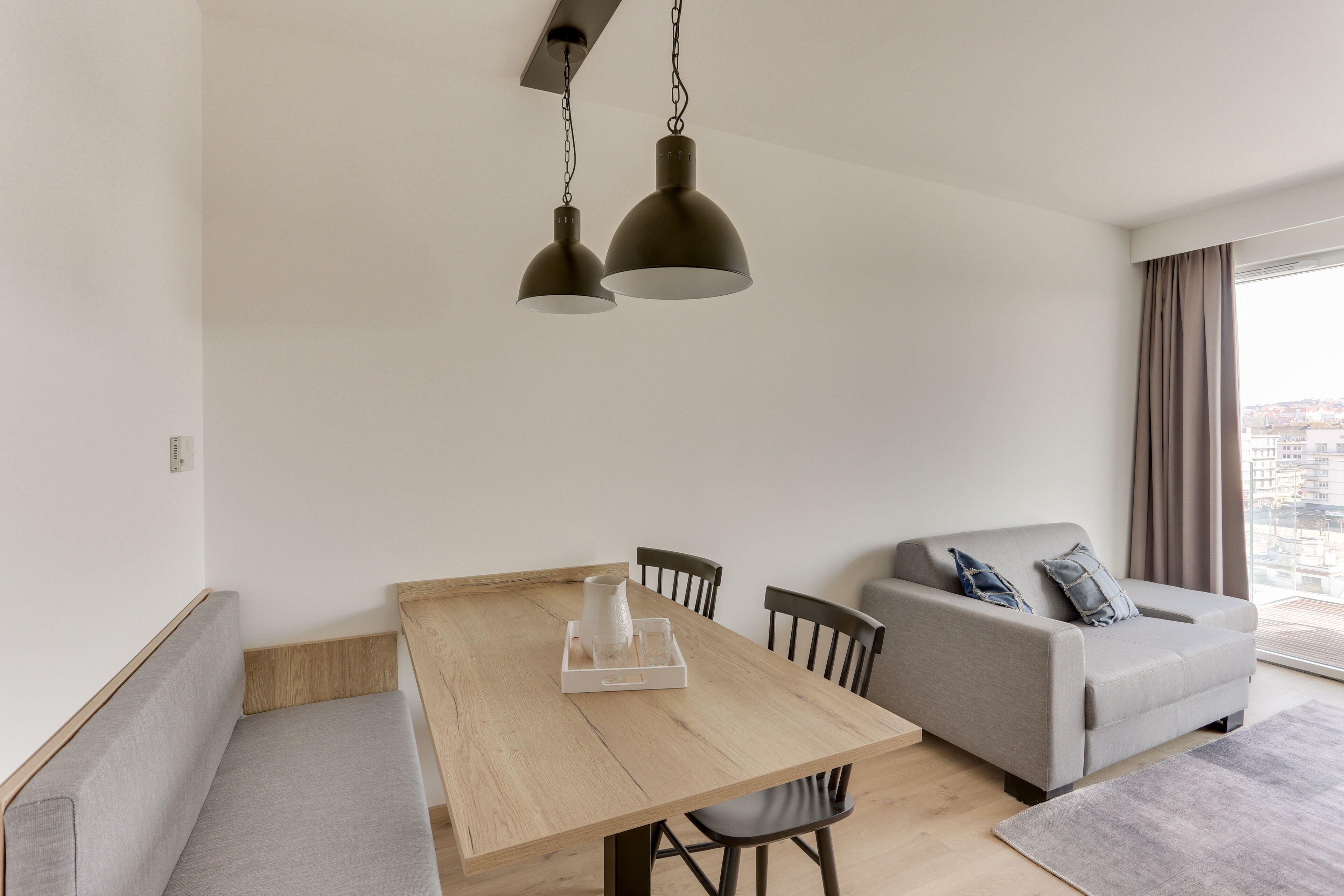 A modern living room with a wooden dining table, grey sofa, and two pendant lights.