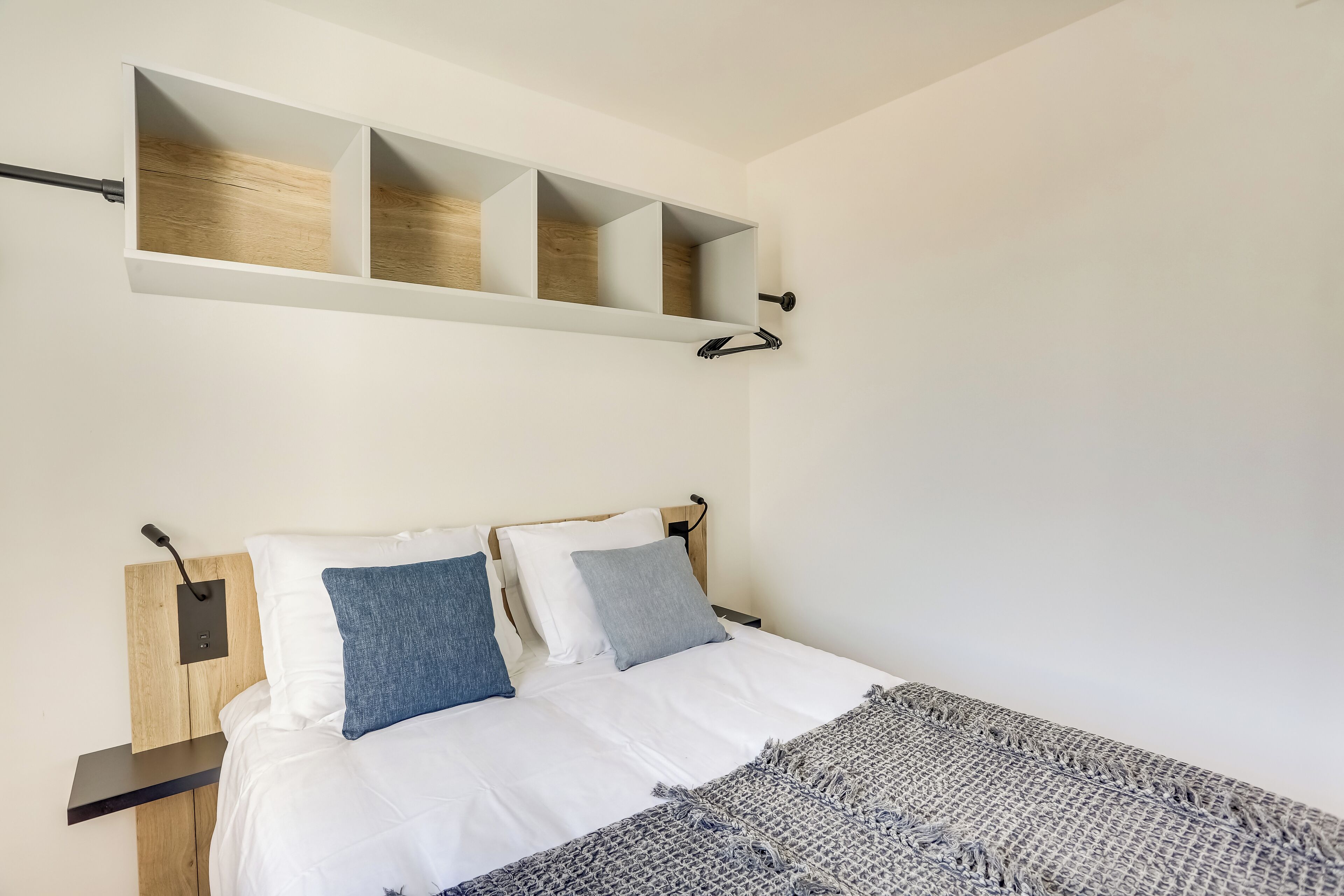 Un dormitorio con cama, dos almohadas, una colcha, una mesita de noche y un estante de pared con tres compartimentos.