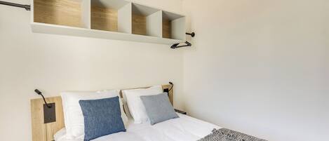 Un dormitorio con cama, dos almohadas, una colcha, una mesita de noche y un estante de pared con tres compartimentos.