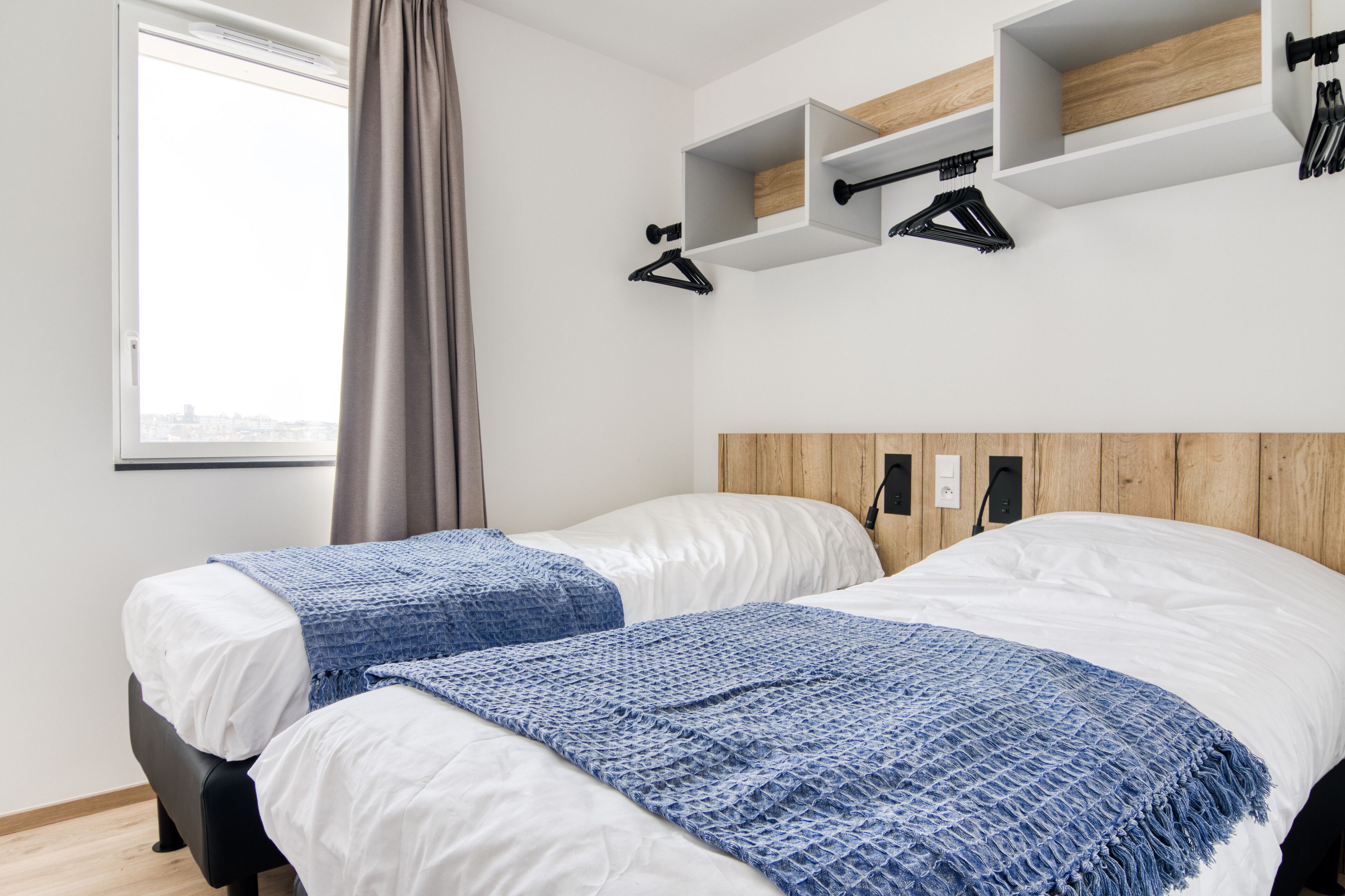A hotel room with two beds, a window with curtains, and built-in wooden shelves above the beds.