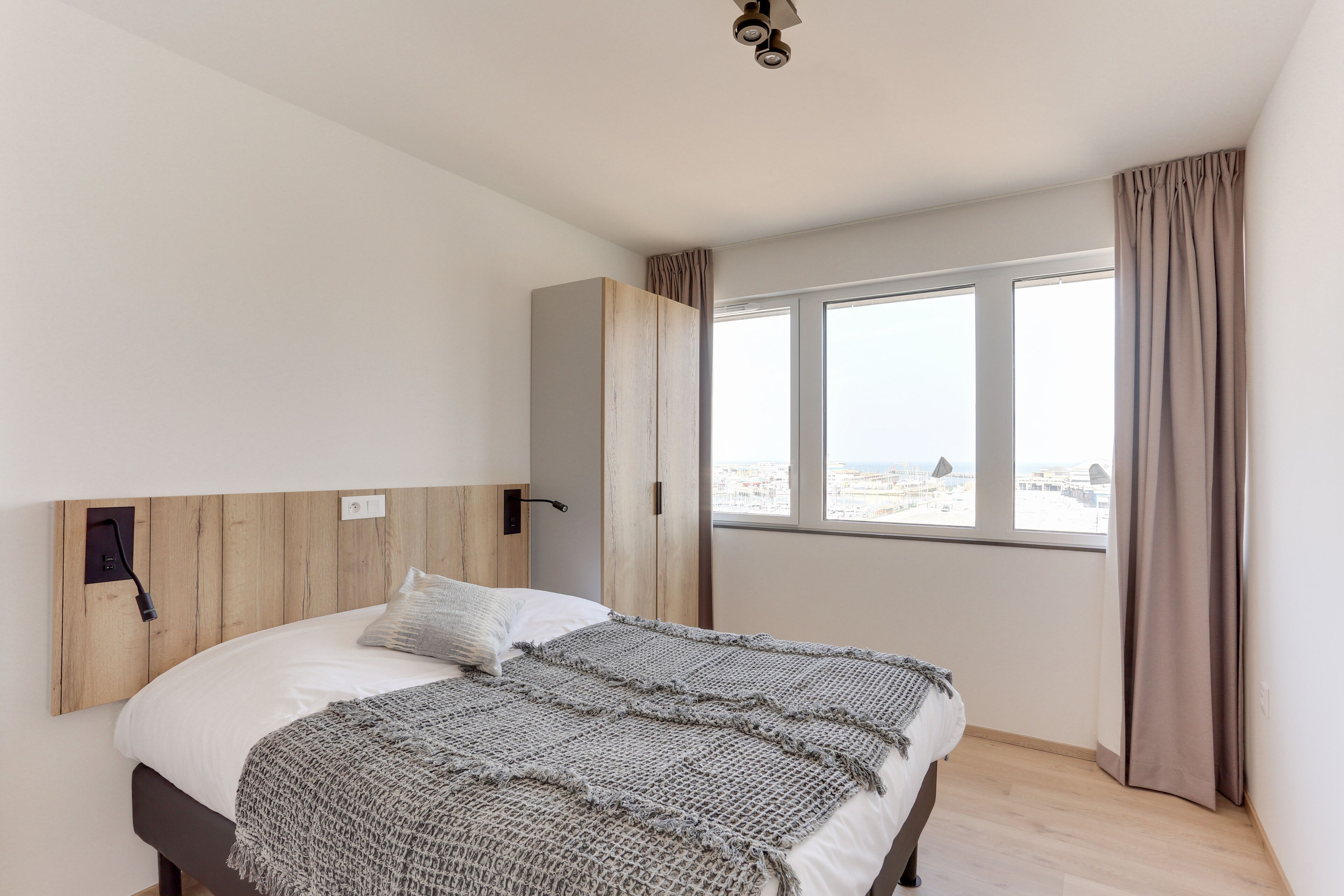 A bedroom with a large bed, a wooden headboard, a window with curtains, and a city view.
