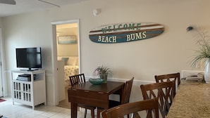 Un salon avec un téléviseur posé sur un meuble, une table à manger avec des chaises, une pancarte indiquant « Bienvenue, les amoureux de la plage », et une plante.