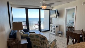 Une chambre avec vue sur la plage, un ventilateur de plafond, un téléviseur fixé au mur, une table à manger avec des chaises et un canapé agrémenté de coussins aux motifs marins.
