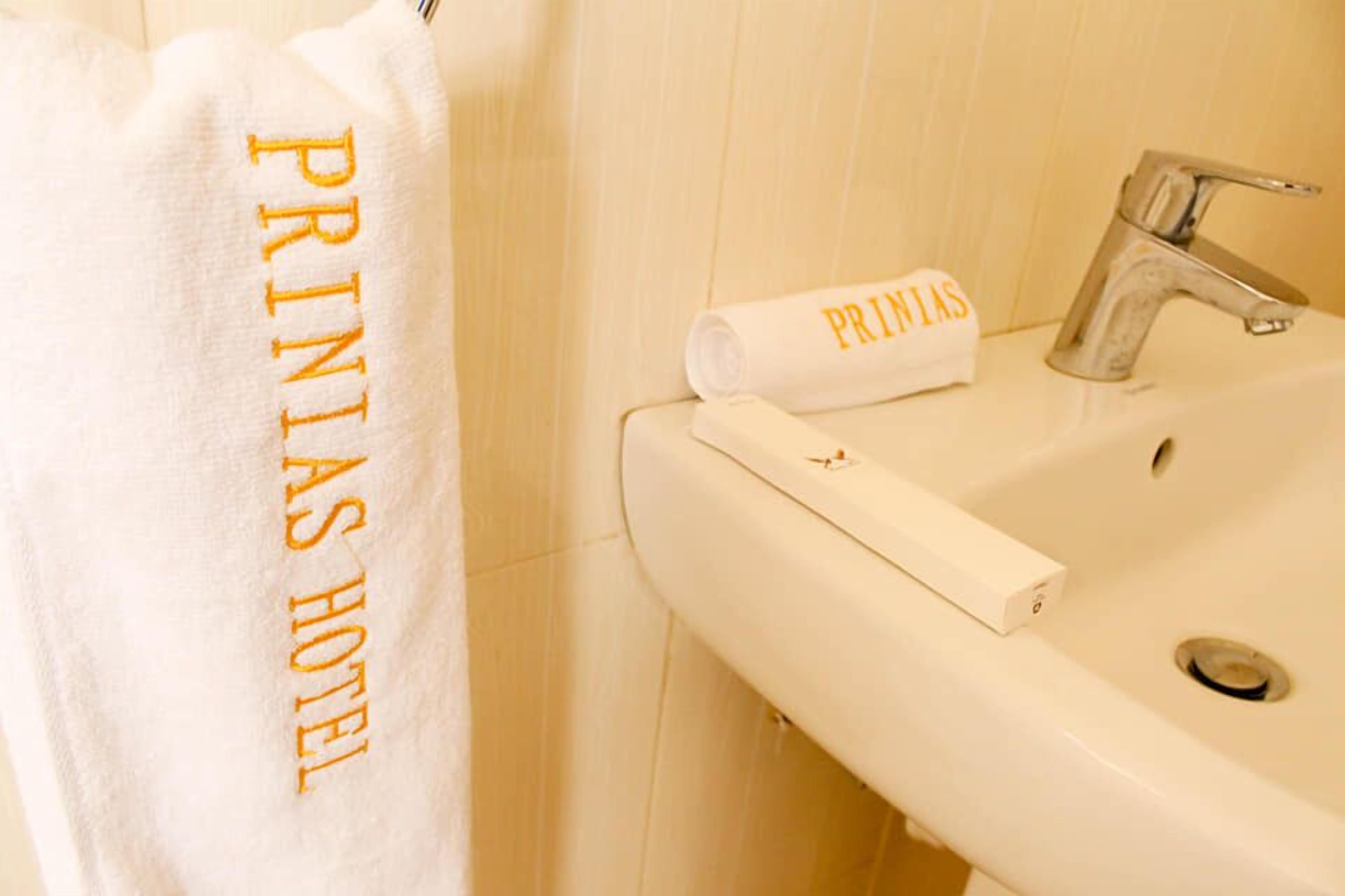 Un baño con un lavamanos blanco, un espejo y un perchero con una toalla que dice "PRINIAS HOTEL".