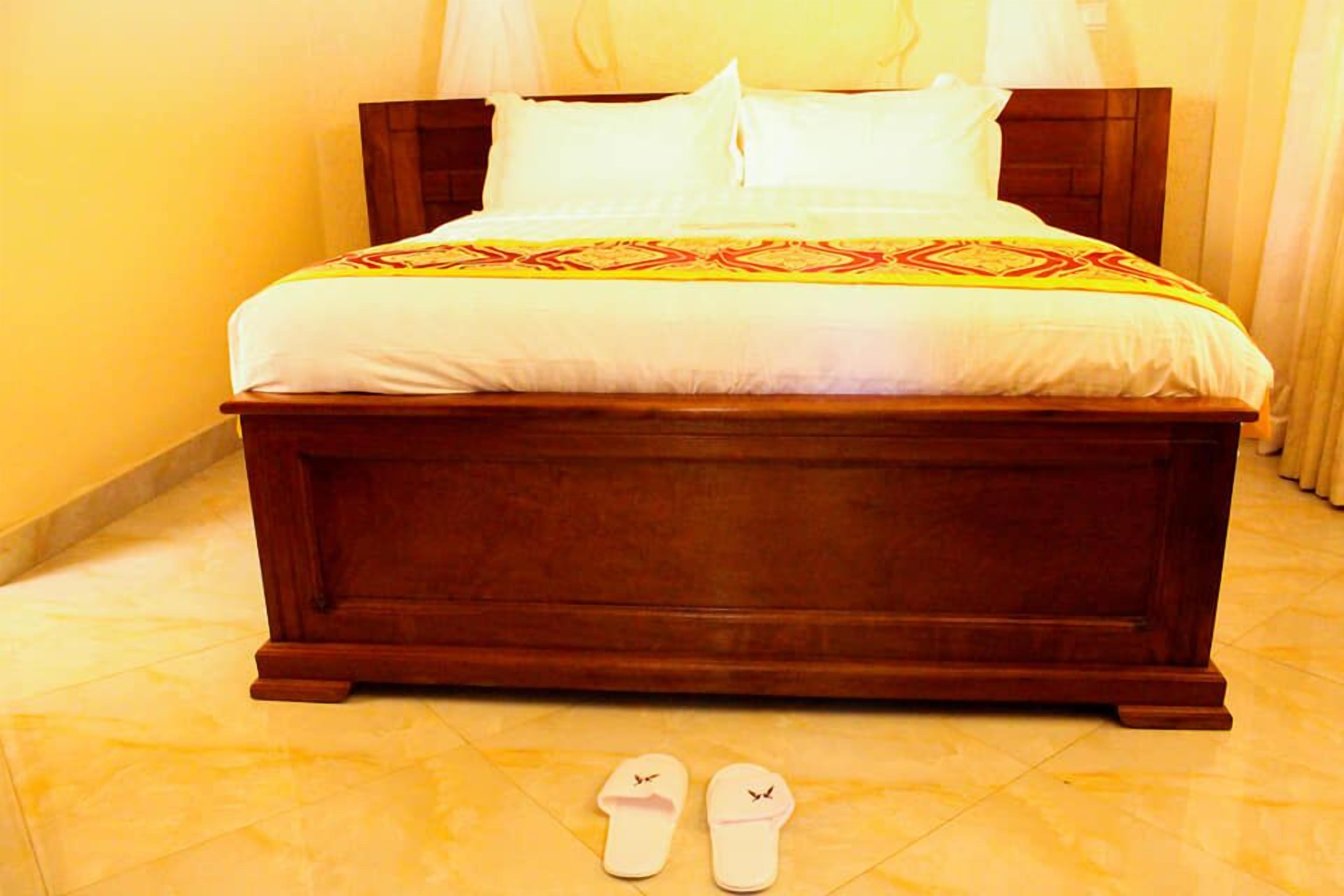 Una cama grande de madera con ropa de cama blanca y una manta estampada, junto a un par de pantuflas blancas en el suelo.
