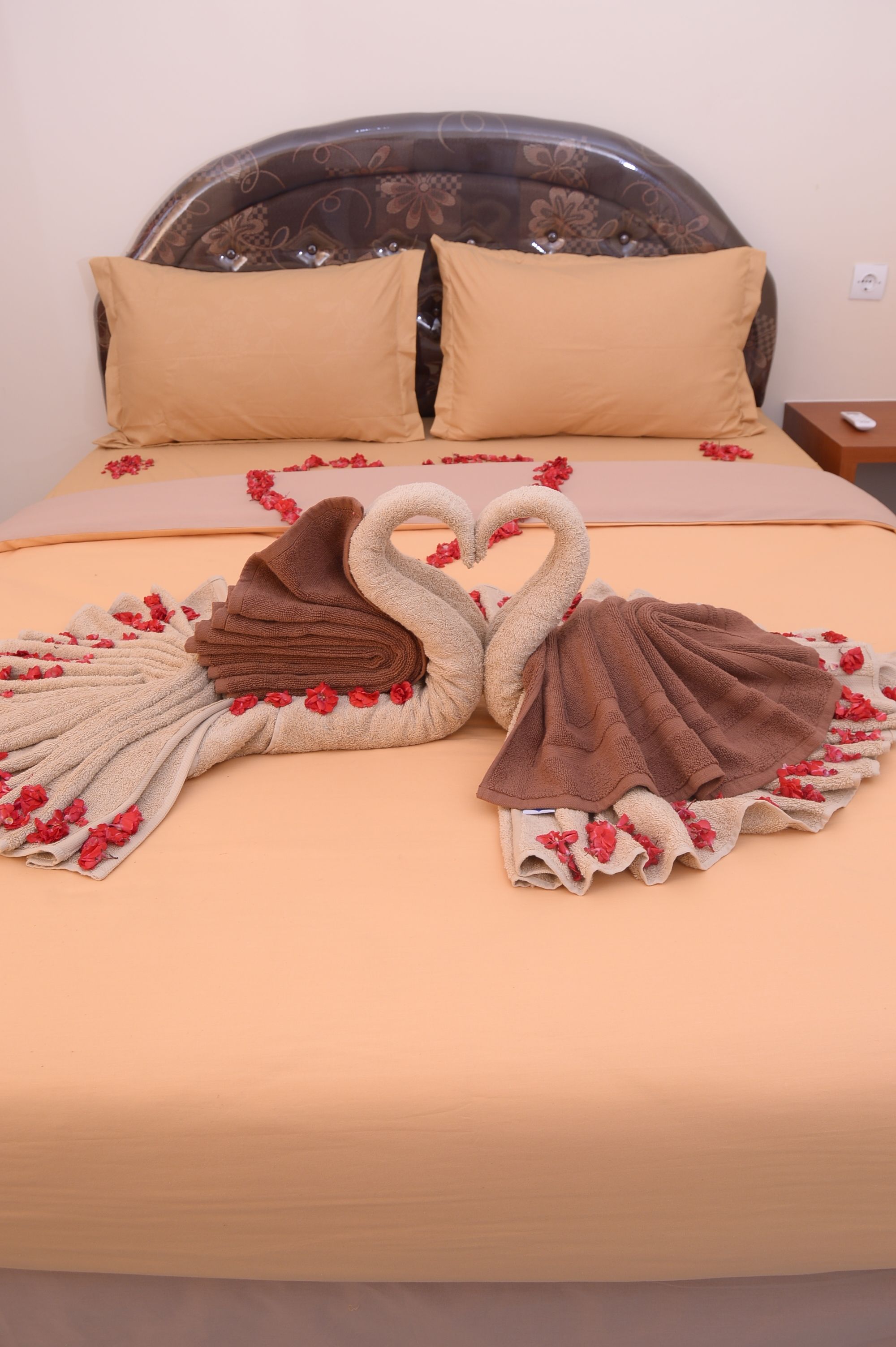 Een bed met een beige deken, twee handdoeken in de vorm van zwanen en rode bloemen.