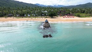 A beach with clear turquoise water, a rock formation, and a resort area with umbrellas and palm trees. - Nacpan Beach Glamping Managed by H Hospitality (El Nido)