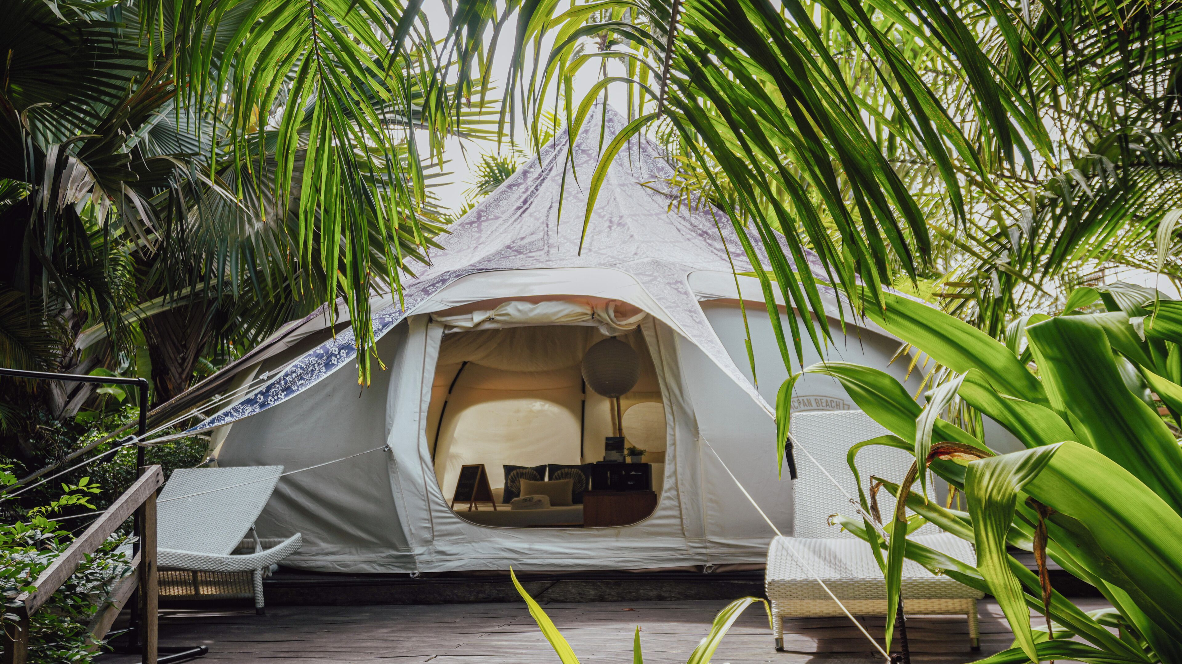 A tent with a bed, a table, and a chair inside, surrounded by lush greenery.
