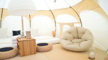 Une pièce confortable aménagée comme une tente, avec un lit, un fauteuil rond en forme de sac de haricots, une table d’appoint en bois sur laquelle se trouvent une bouteille et un calendrier, et une lampe.