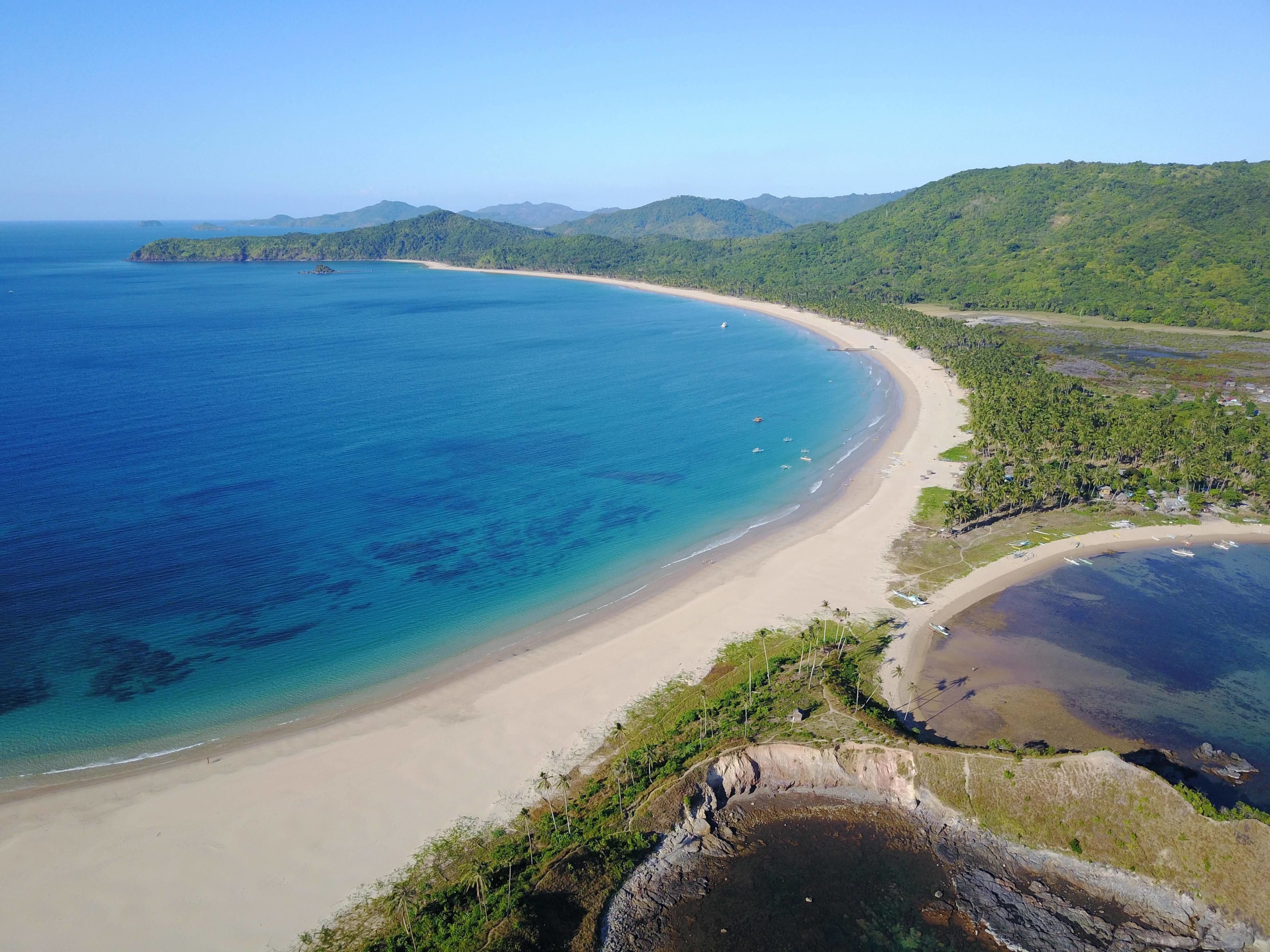 Paisaje costero con playa arenosa, aguas azules cristalinas y colinas verdes exuberantes al fondo.
