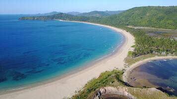 Paisaje costero con playa arenosa, aguas azules cristalinas y colinas verdes exuberantes al fondo.