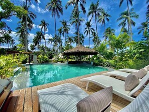 A pool area with lounge chairs, a thatched-roof structure, and tall palm trees.