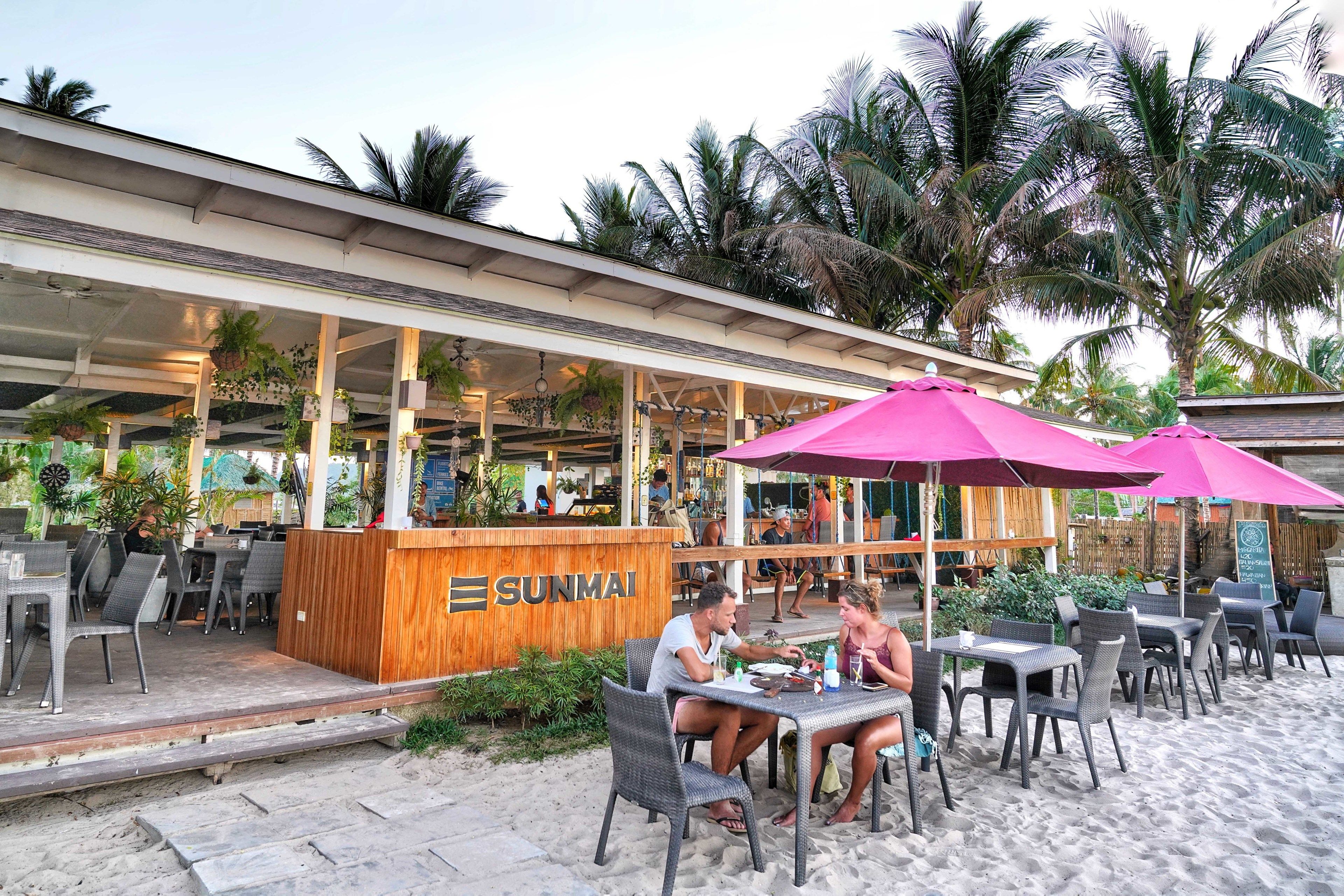 An outdoor seating area with tables and chairs, a wooden counter with the name "SUNMAI", and palm trees in the background.