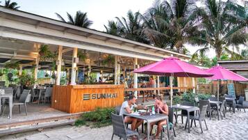 An outdoor seating area with tables and chairs, a wooden counter with the name "SUNMAI", and palm trees in the background.