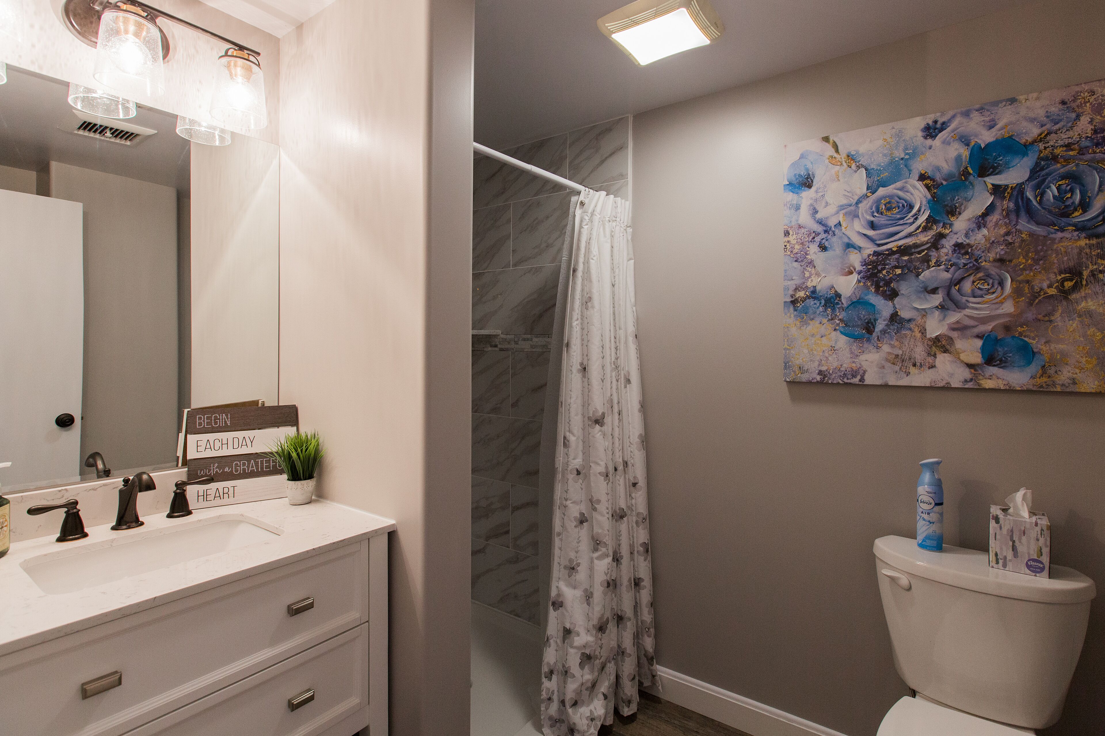 Une salle de bain avec un lavabo blanc, un miroir, un rideau de douche et des toilettes. Il y a un panneau sur le lavabo et un tableau au mur.