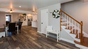 Een ruime woonkamer met houten vloer, een eettafel met stoelen, een keuken op de achtergrond en een trap met een witte balustrade.