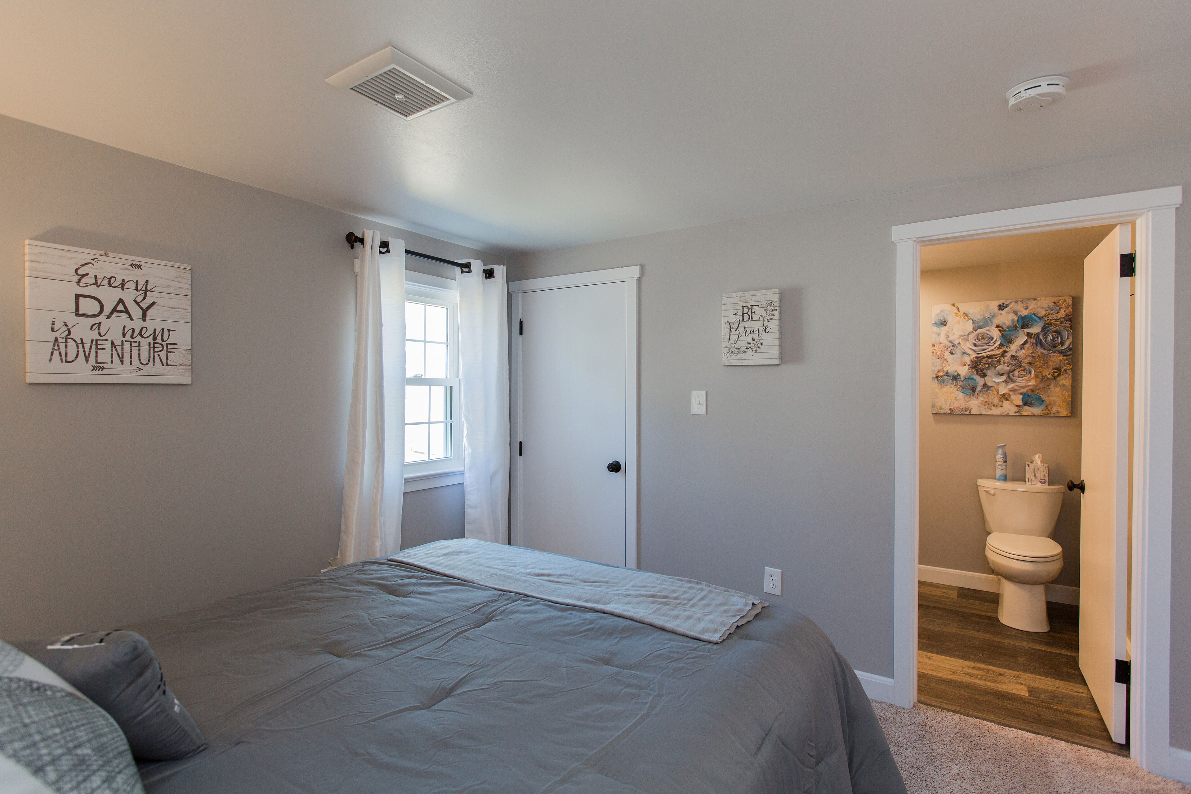 A bedroom with a bed, a window with curtains, a door, and a bathroom visible through an open door.