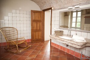 A bathroom with a wooden door, a wicker chair, a sink with a mirror, and a tiled floor. - Cascina San Giorgio (Valenza)