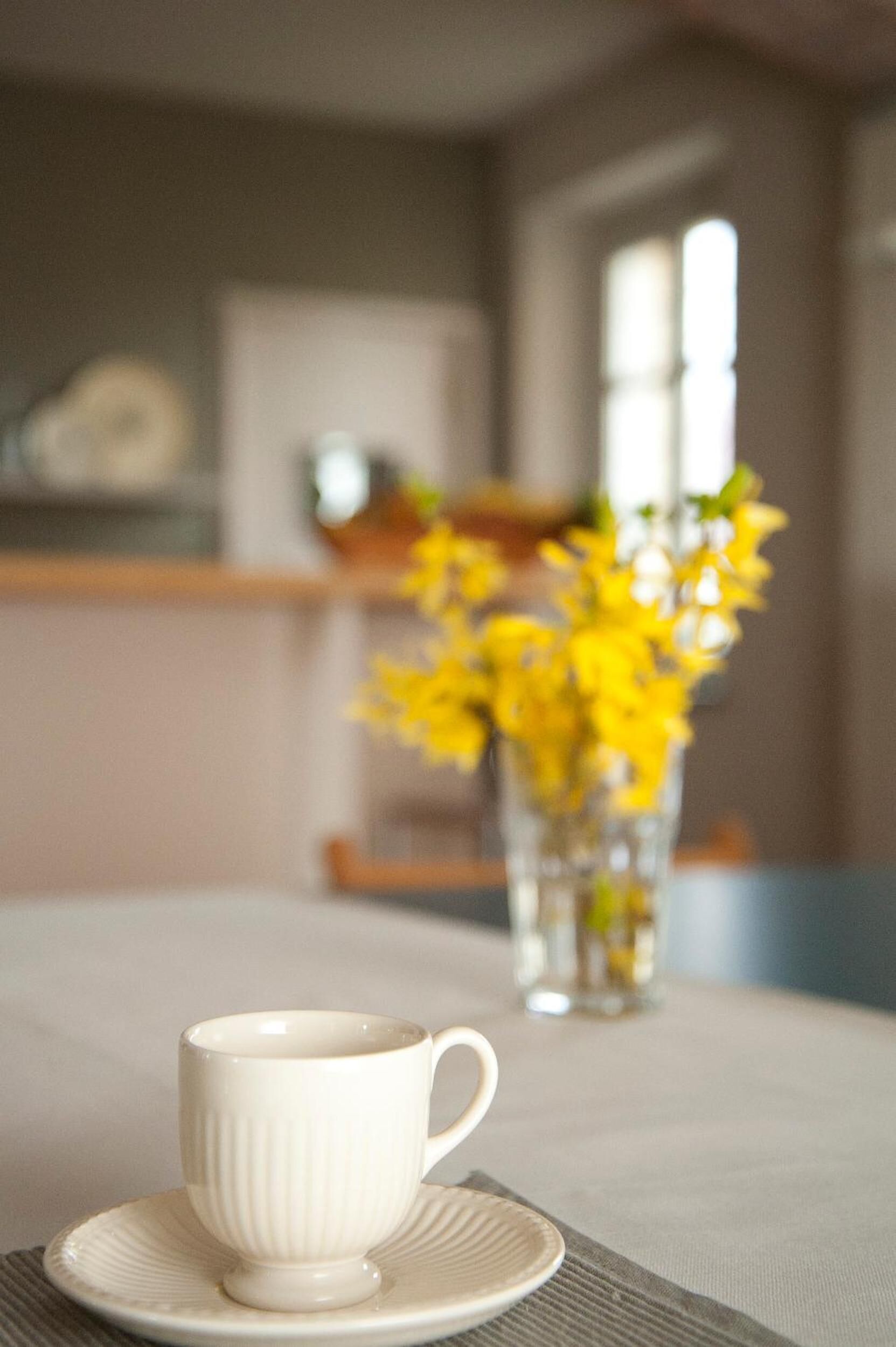 A cup on a saucer on a table with a vase of yellow flowers in the background.