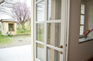 A view through a partially open door with a window, a small potted plant, and a tree with pink blossoms. - Cascina San Giorgio (Valenza)