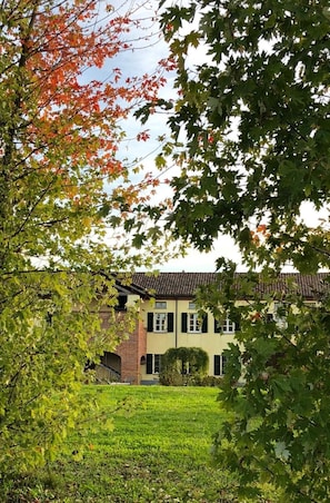 A building with a brick facade and multiple windows, surrounded by lush greenery and trees with autumn leaves. - Cascina San Giorgio (Valenza)