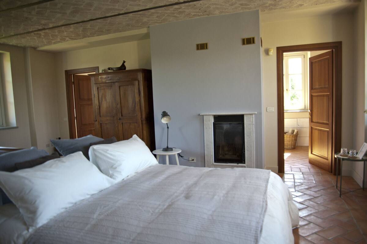 A bedroom with a large bed, a small table with a lamp, a wooden wardrobe, and a fireplace.
