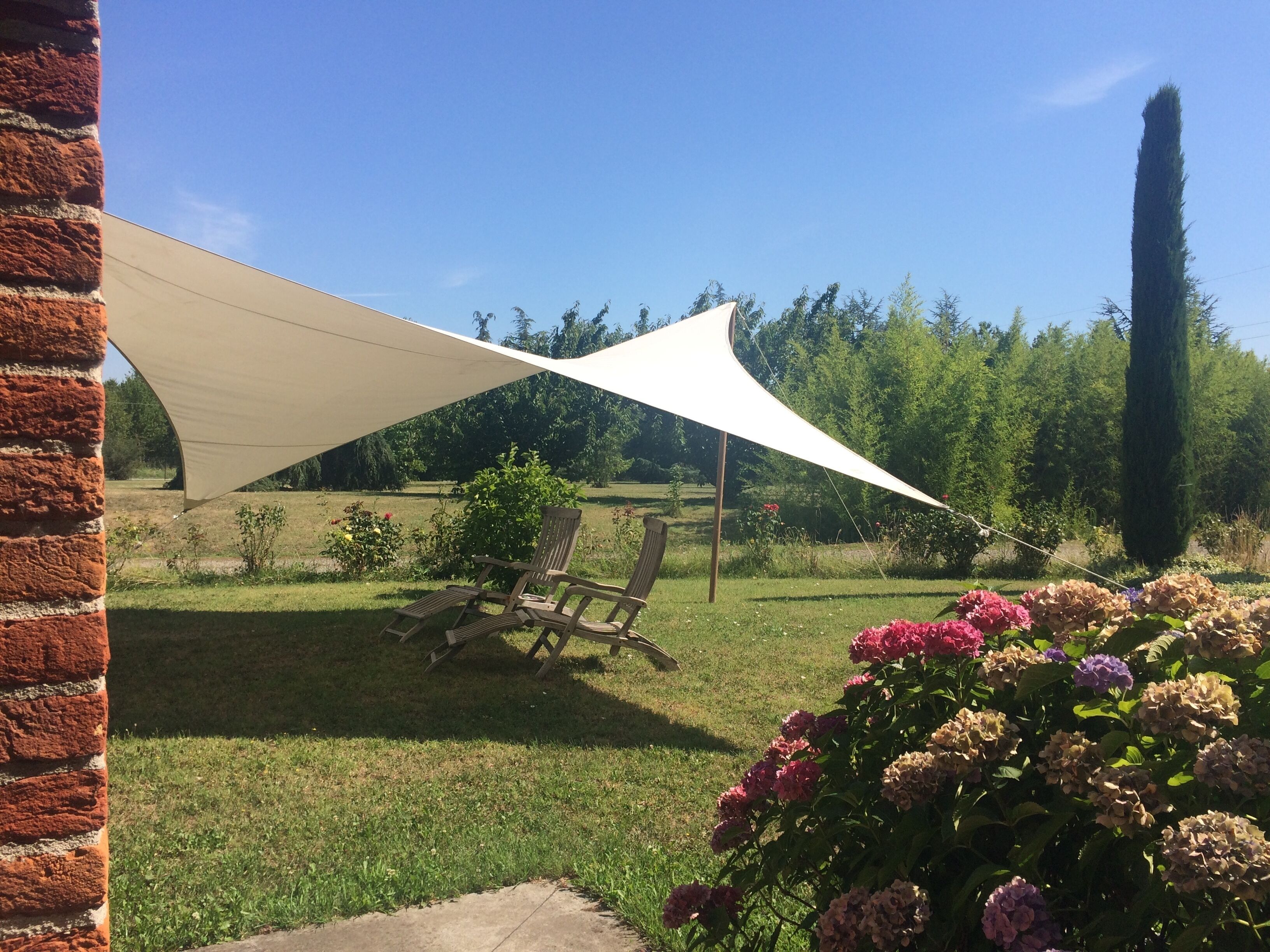 Un espacio al aire libre con una vela de sombra grande, una silla de madera y un macizo de flores.