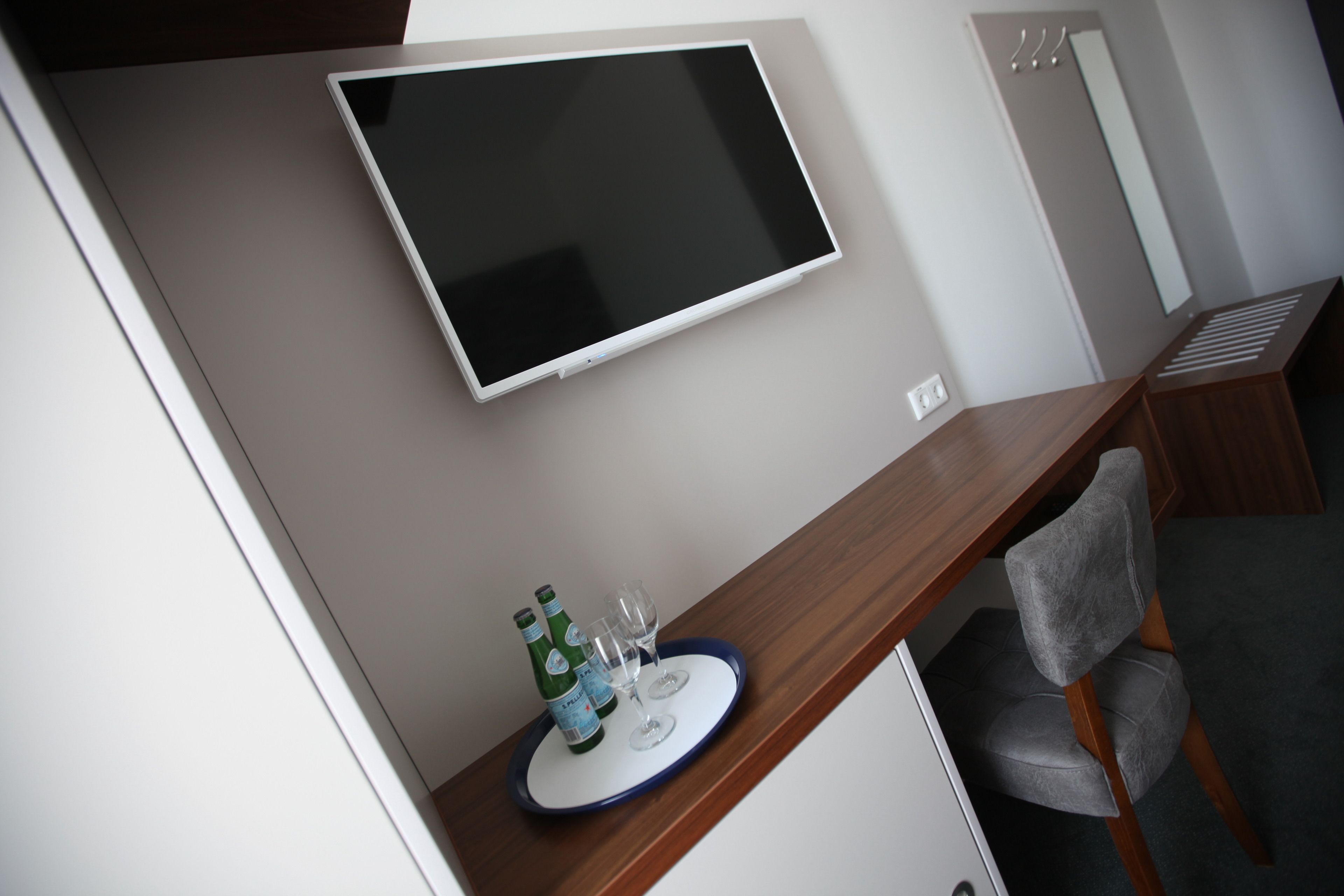 Una habitación moderna con un televisor montado, un escritorio de madera, una silla gris y una mesita con una bandeja que contiene una botella y vasos.