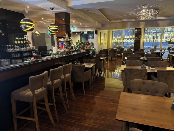 A restaurant with a bar area, wooden tables and chairs, and large windows with a view. - Hotel Starton am Ingolstadt Village (Ingolstadt)
