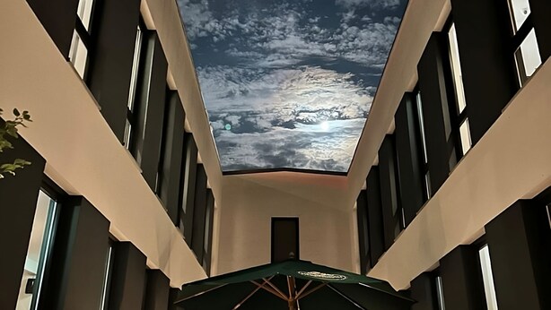 A modern hotel interior with a view of the sky through a skylight, a dining area with tables and chairs, and a green umbrella. - Hotel Starton am Ingolstadt Village (Ingolstadt)