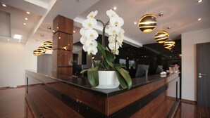 A modern reception desk with a potted orchid, wooden paneling, and contemporary pendant lighting. - Hotel Starton am Ingolstadt Village (Ingolstadt)
