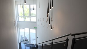A modern staircase with a glass railing, leading to a bright area with large windows and hanging pendant lights. - Hotel Starton am Ingolstadt Village (Ingolstadt)