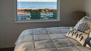 Een slaapkamer met een groot bed, een plafondventilator en een raam met uitzicht op het strand en stoelen buiten.