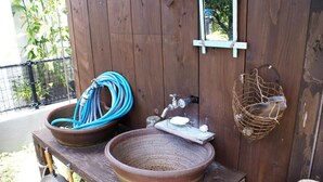 Un baño al aire libre rústico con dos lavamanos grandes de cerámica, una manguera, un jabonero y un estante de madera con una cesta.