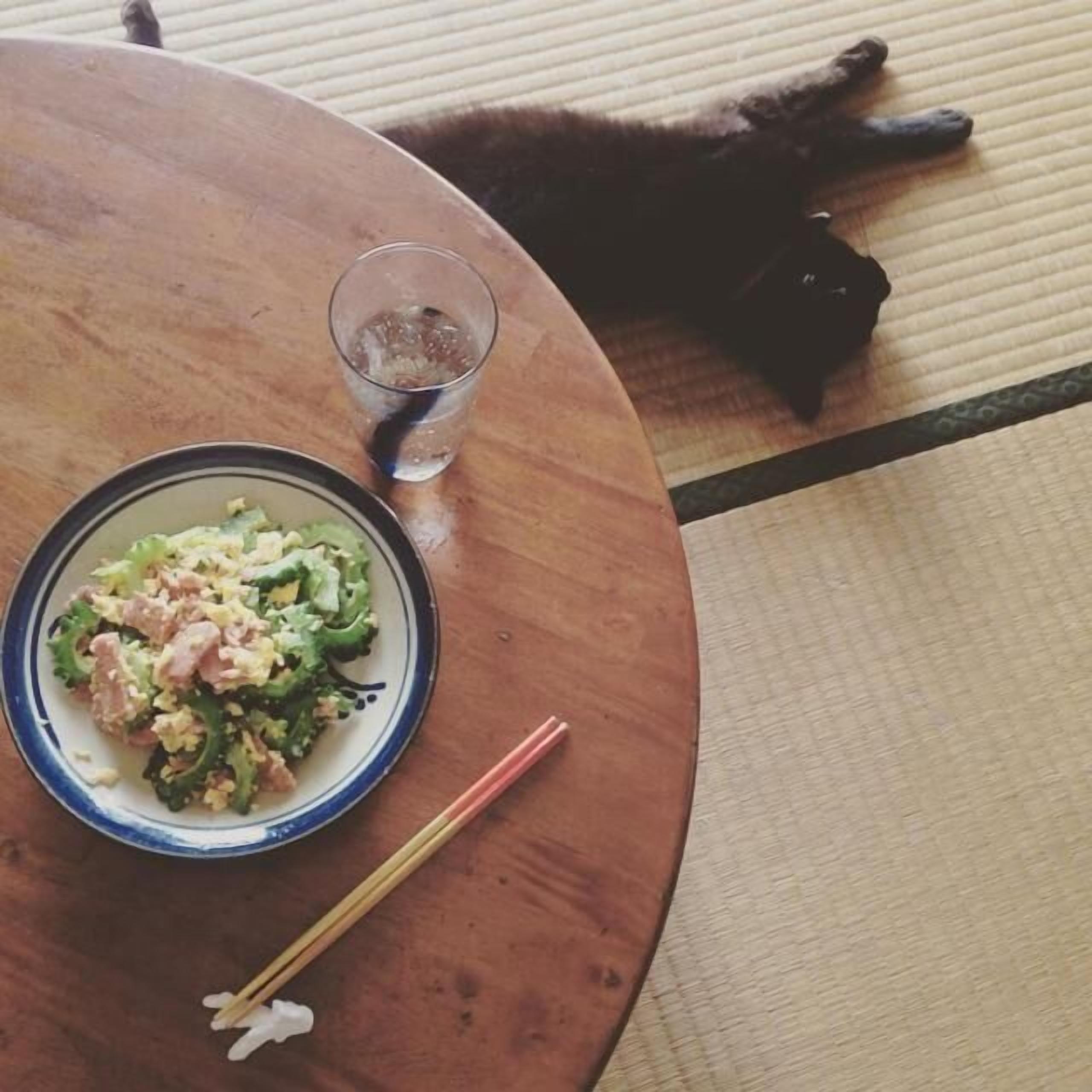 A round wooden table with a plate of food and chopsticks, a glass of water, and a black cat lying on a mat.