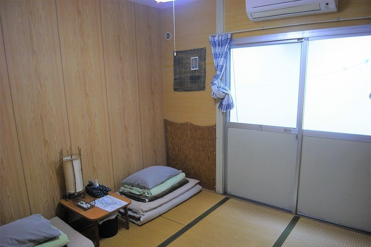 Une chambre avec deux lits, une petite table avec une lampe, une fenêtre avec un rideau et un climatiseur fixé au mur.