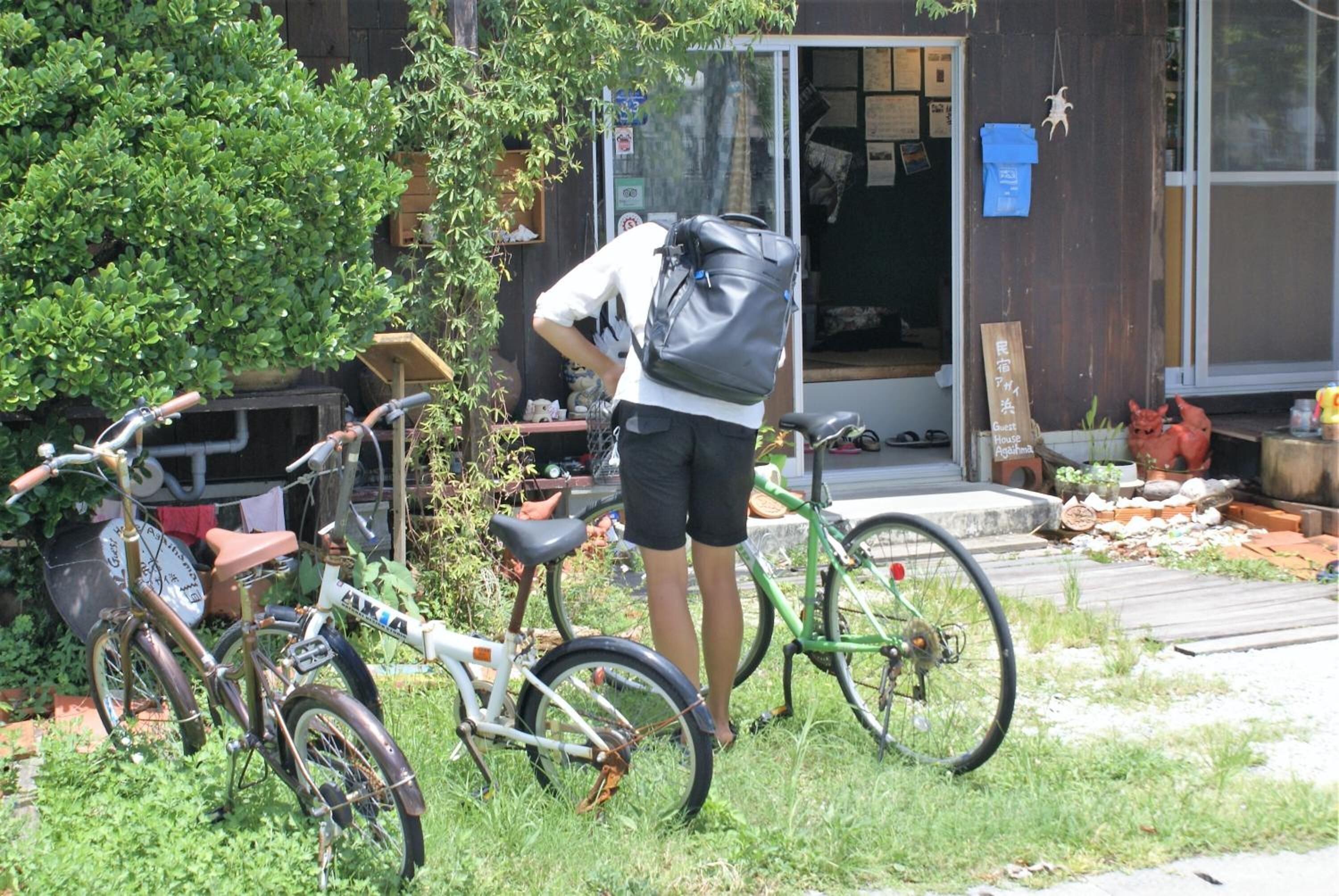 Eine Person steht neben einem Fahrrad in einem Garten, im Hintergrund ist ein Gebäude zu sehen.