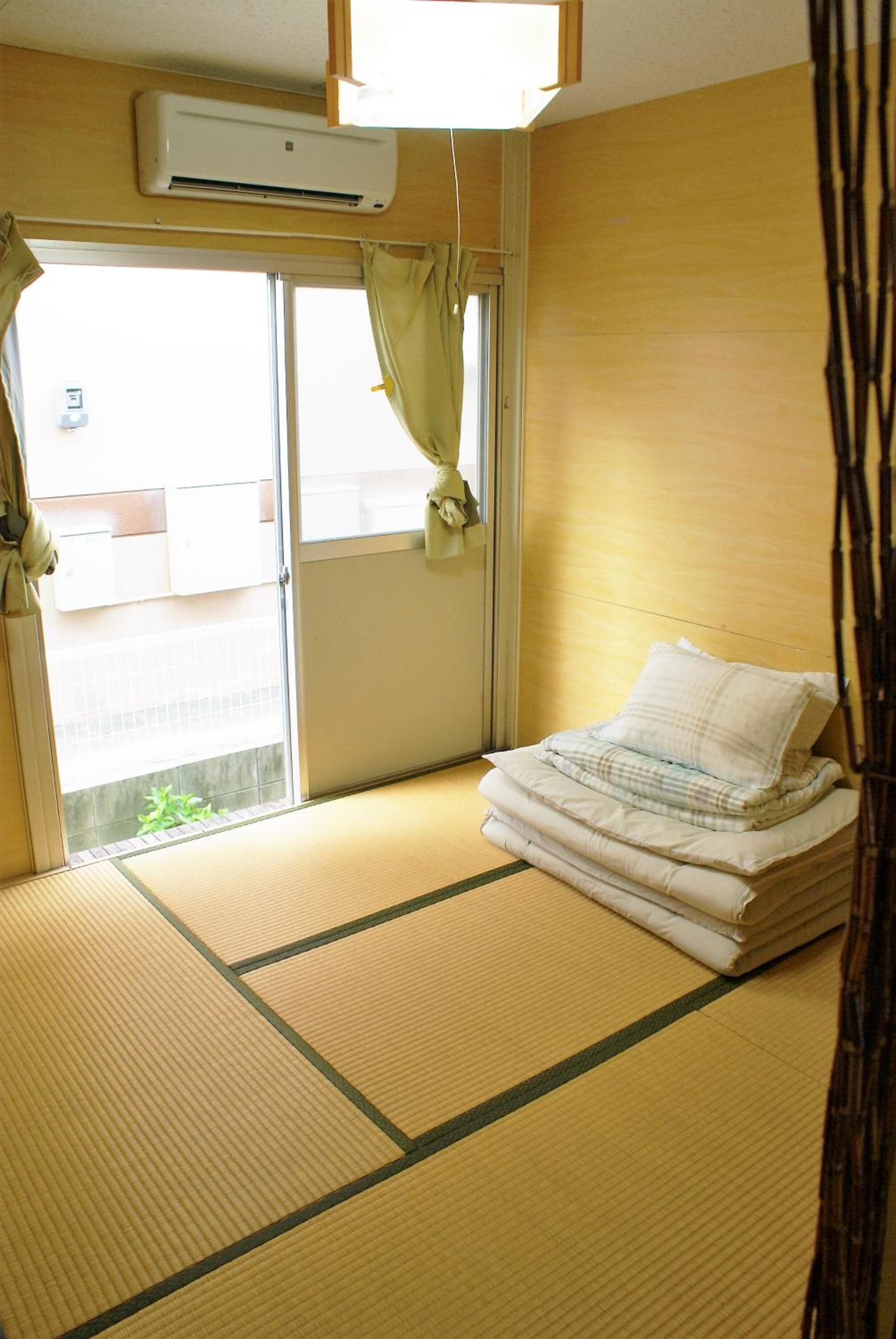 A room with tatami flooring, a bed with a white mattress and pillow, an air conditioning unit, and a sliding door with a view of the outside.