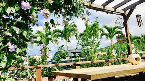 Una pérgola de madera con una mesa y bancos, rodeada de abundante follaje y plantas en flor.