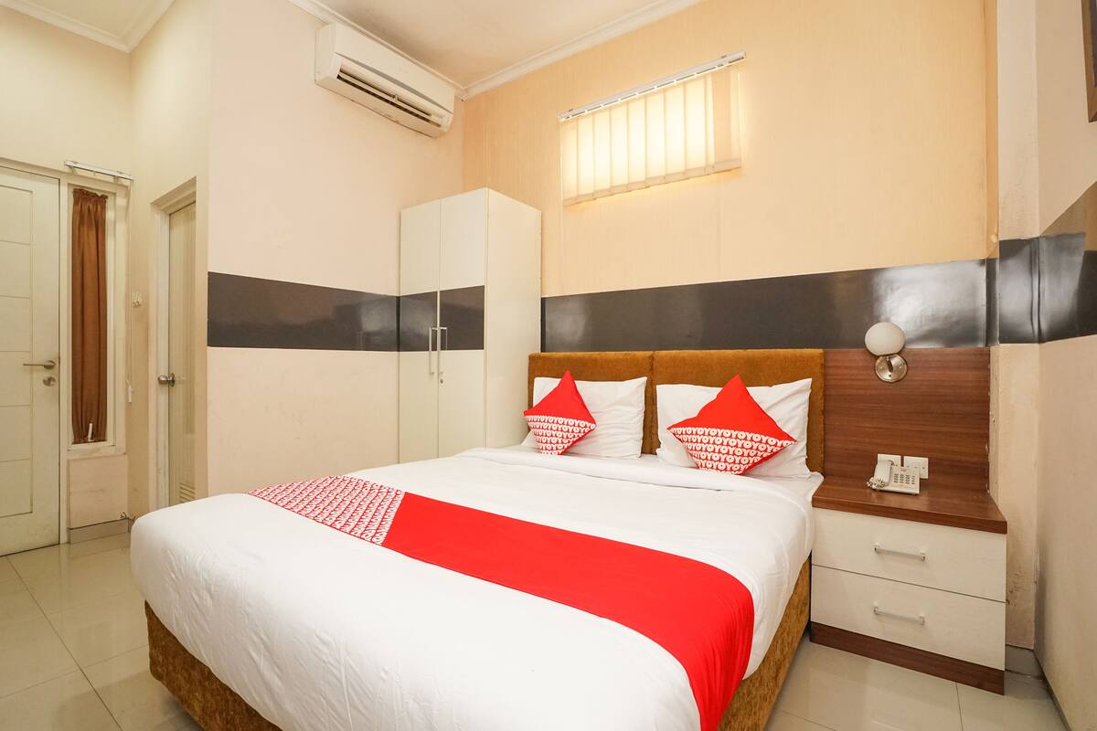 A hotel room with a bed, a wardrobe, a bedside table with a phone, and a window with blinds.