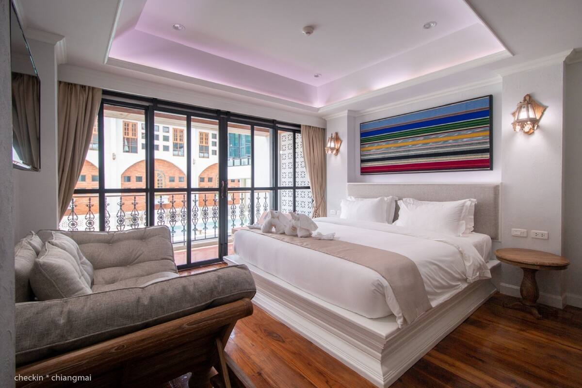 A modern hotel room with a large bed, a sofa, and a view of a building through large windows.