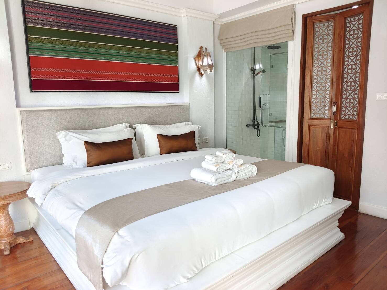 A neatly made bed with white linens and brown pillows, a glass shower door, and a wooden door with decorative glass panels.